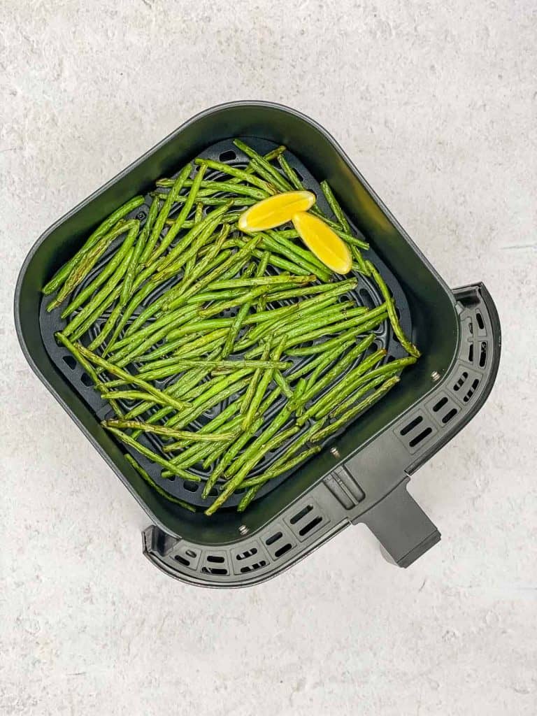 An air fryer basket with cooked green beans and a lemon wedge.