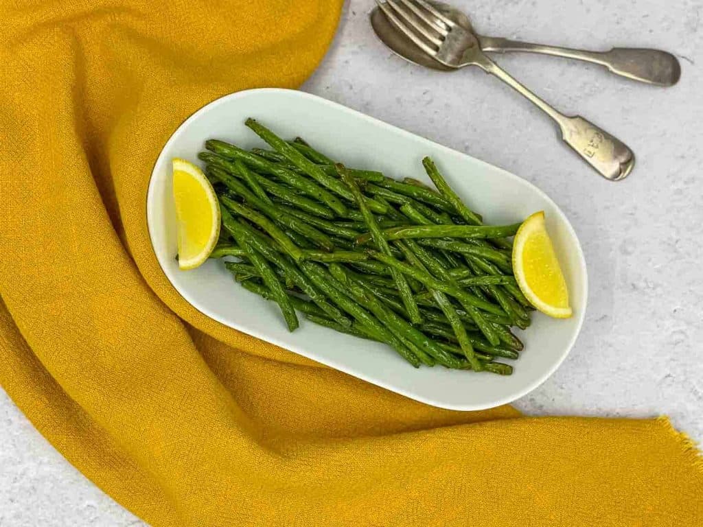 A serving dish of air fryer green beans with lemon wedges and a spoon and fork.