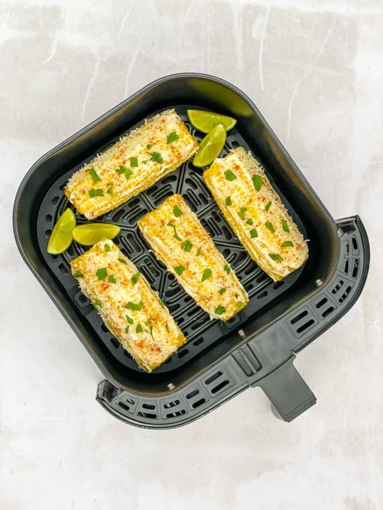 Four corn on the cobs Mexican style, with lime wedges in the air fryer basket.