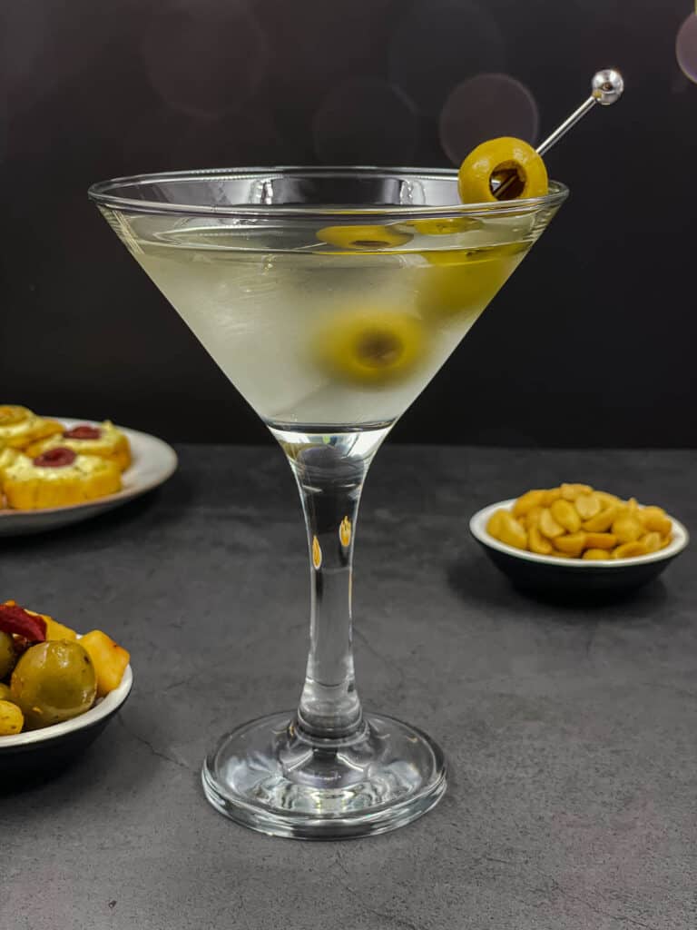 Homemade dirty martini in a cocktail glass with a green olive garnish, and nuts and olive snacks.
