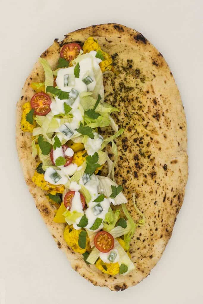 A naan bread with aloo gobi, raita, tomatoes, and lettuce on one half.