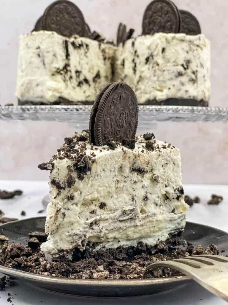A delicious slice of no bake Oreo cheesecake on a plate, with a whole cheesecake behind it on a cake stand.