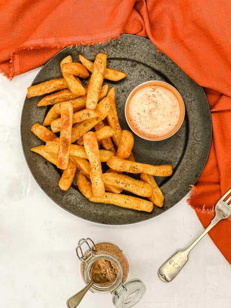 A plate of peri peri fries on a plate, with a bowl of dipping sauce, a fork, and a jar of peri peri seasoning.