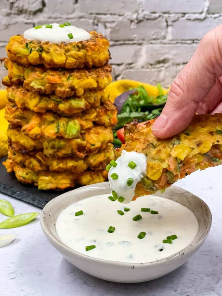 Someone dipping half a vegetable fritter in sour cream and chives with chopped chives on top, a stack of vegetable fritters behind.