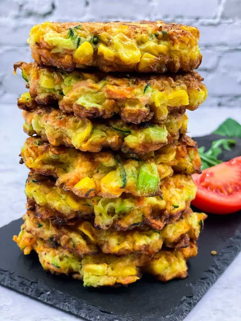 Vegetable fritters stacked on a slate board with tomato and rocket in the background.