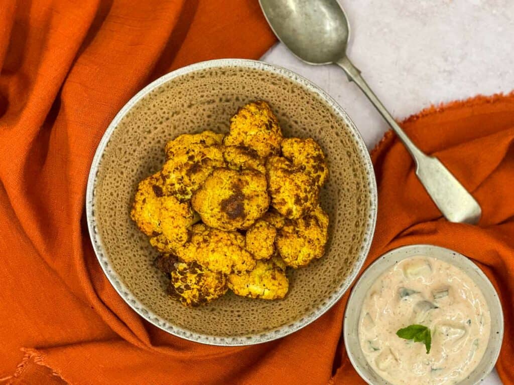 Crispy air fryer cauliflower in a bowl with a serving bowl and a dipping sauce.