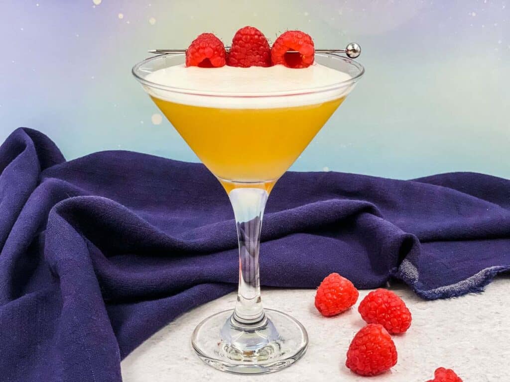 A martini glass filled with a French martini cocktail, with fresh raspberries as garnish and scattered around on the surface.