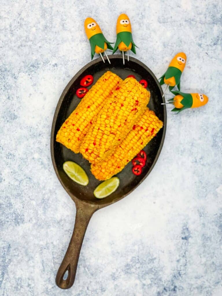 A skillet with chili and lime corn on the cob with lime wedges, chili peppers, and corn holders.