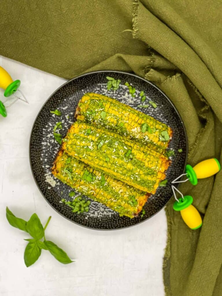 A plate with sweet corn on the cob with pesto butter, Parmesan and chopped basil leaves, corn holders and some fresh basil leaves.