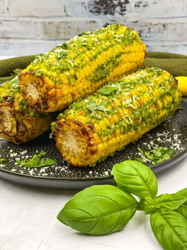Three corn on the cobs with pesto butter and Parmesan on them, basil leaves on the side.