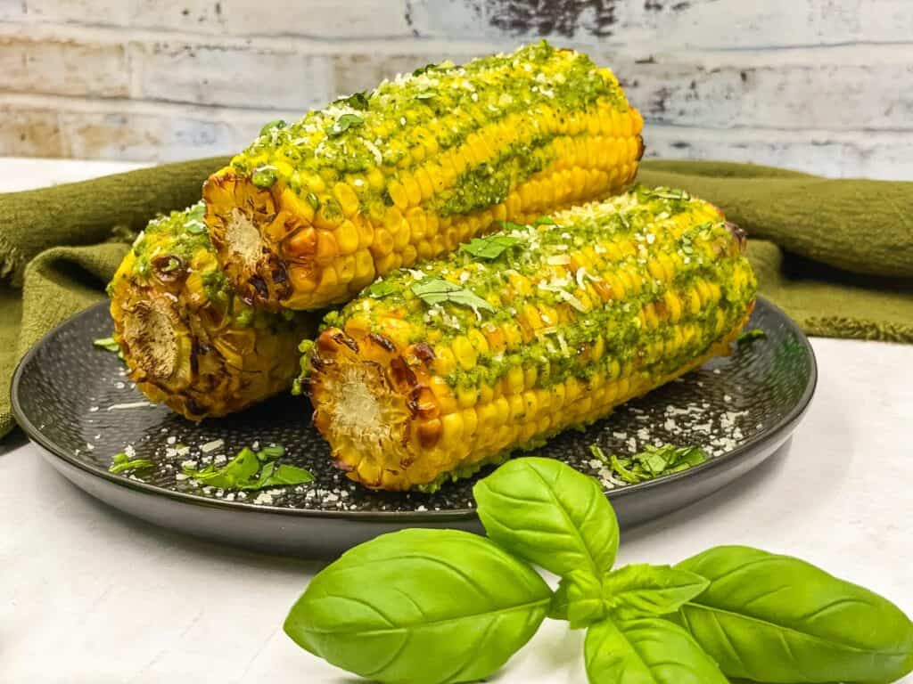 Pesto corn on the cob on a plate with fresh basil leaves by them.