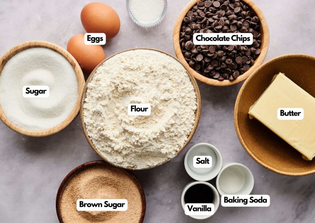 Bowls containing the classic ingredients for chocolate chip cookies—flour, sugar, brown sugar, chocolate chips, butter, eggs, salt, vanilla, and baking soda—are artfully arranged on a light surface. Each ingredient is clearly labeled.