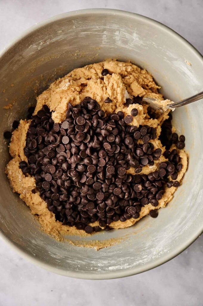 A mixing bowl with cookie dough and a generous amount of chocolate chips, perfect for making delicious chocolate chip cookies, is being stirred with a spoon.