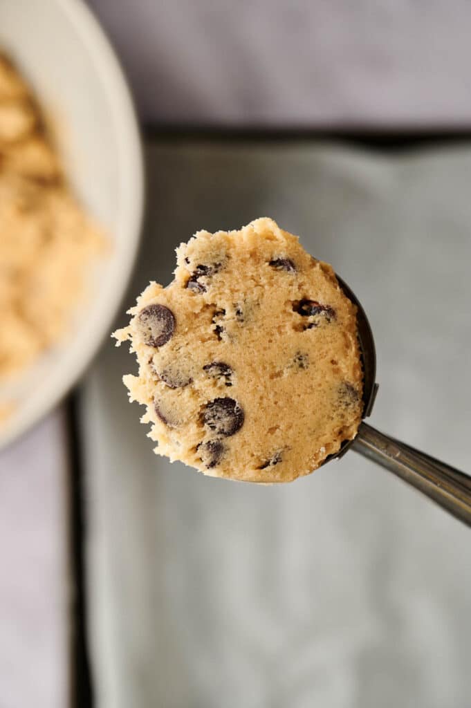 Close-up of a metal scoop holding a portion of chocolate chip cookie dough, with a blurred background featuring a bowl of chocolate chip cookies and a baking sheet.