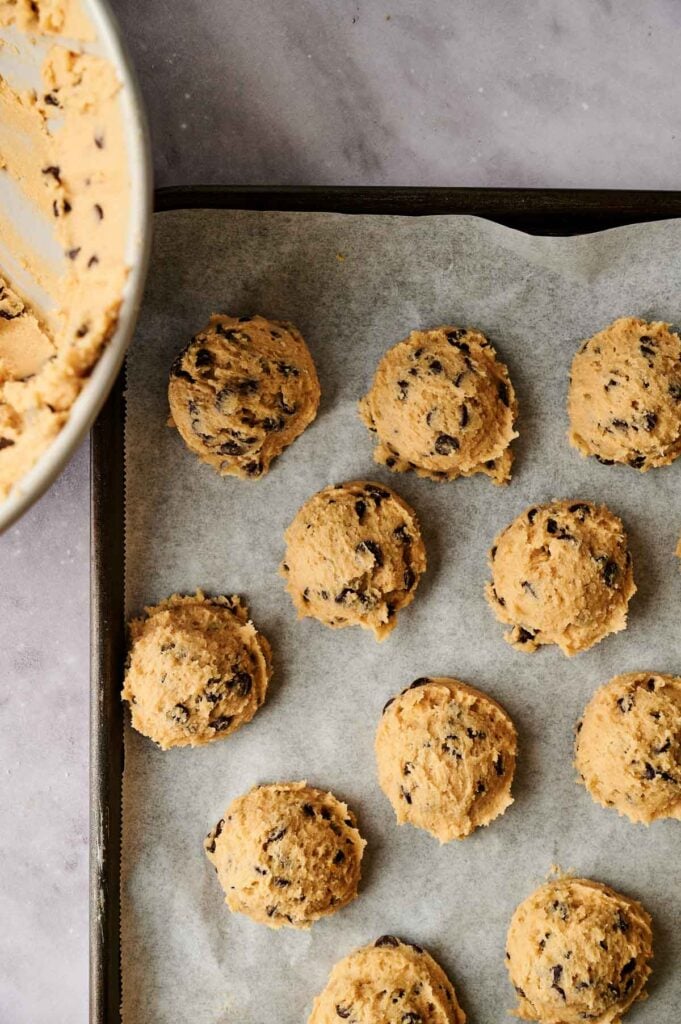 A baking tray lined with parchment paper holds multiple scoops of raw chocolate chip cookie dough, ready to become delicious chocolate chip cookies. A mixing bowl of dough is partially visible on the left side.