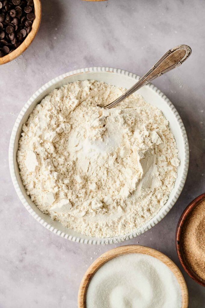 A bowl filled with flour and a spoon inside, surrounded by smaller bowls containing chocolate chips, sugar, and brown sugar on a marble surface—the perfect scene to start baking chocolate chip cookies.