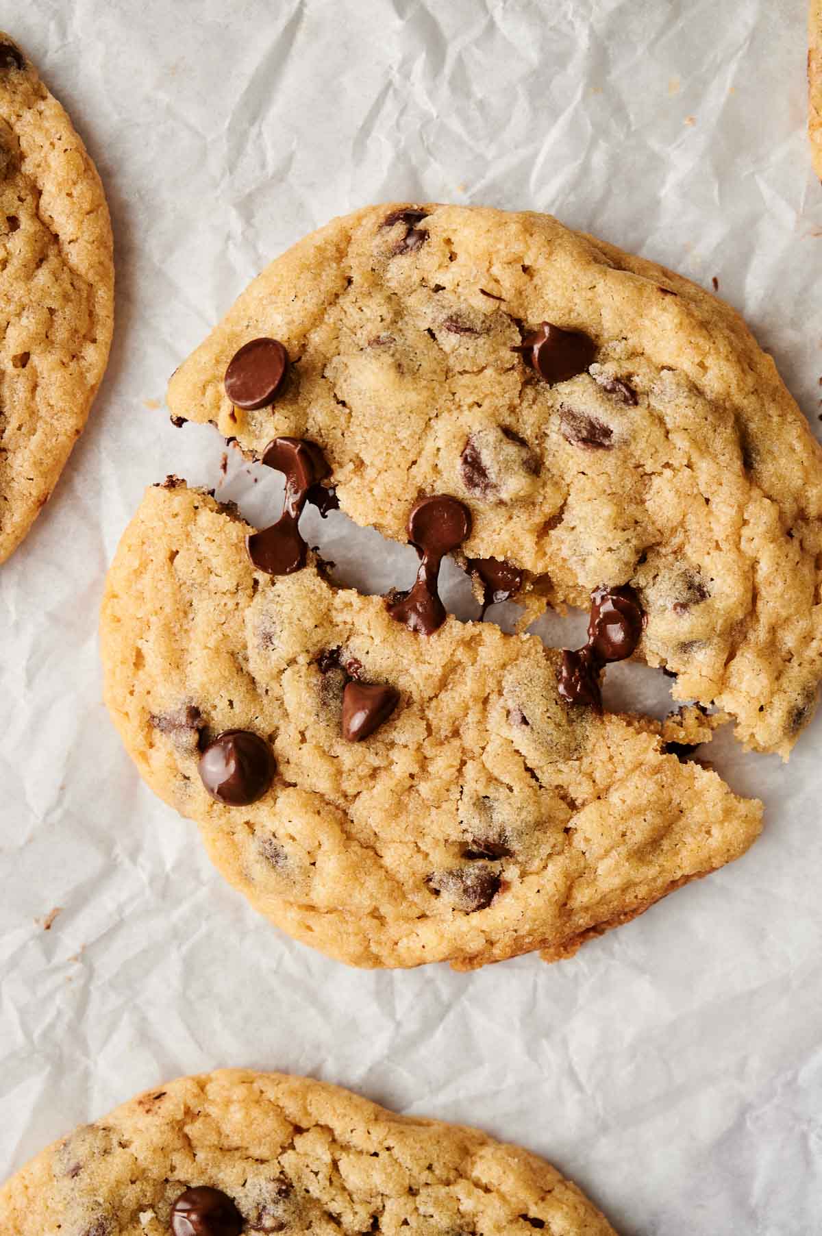 Close-up of chocolate chip cookies, with one broken into two pieces, placed on parchment paper, surrounded by other whole cookies.