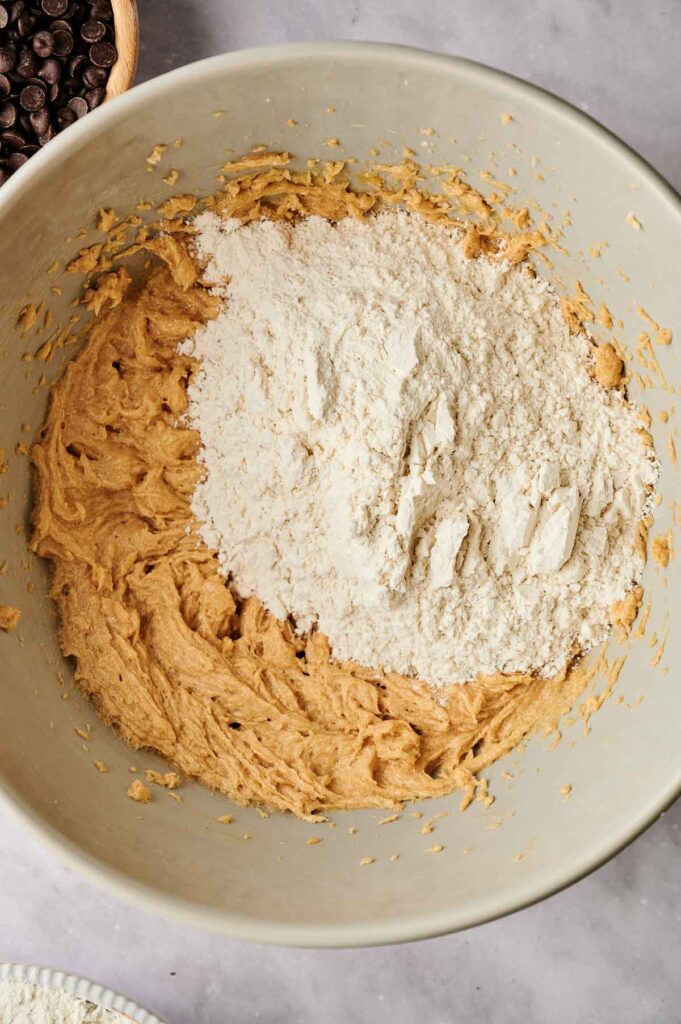 A mixing bowl containing partially mixed chocolate chip cookie dough with a pile of flour on one side.