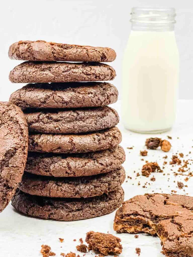 A stack of double chocolate chip cookies, one with a bite out of it, and a jug of milk.