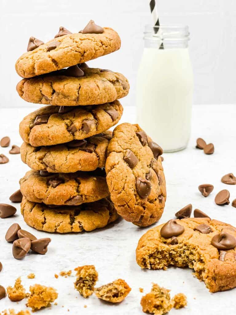 A stack of Peanut butter chocolate chip cookies, with one cookie having a bite taken out of it and a jar of milk.