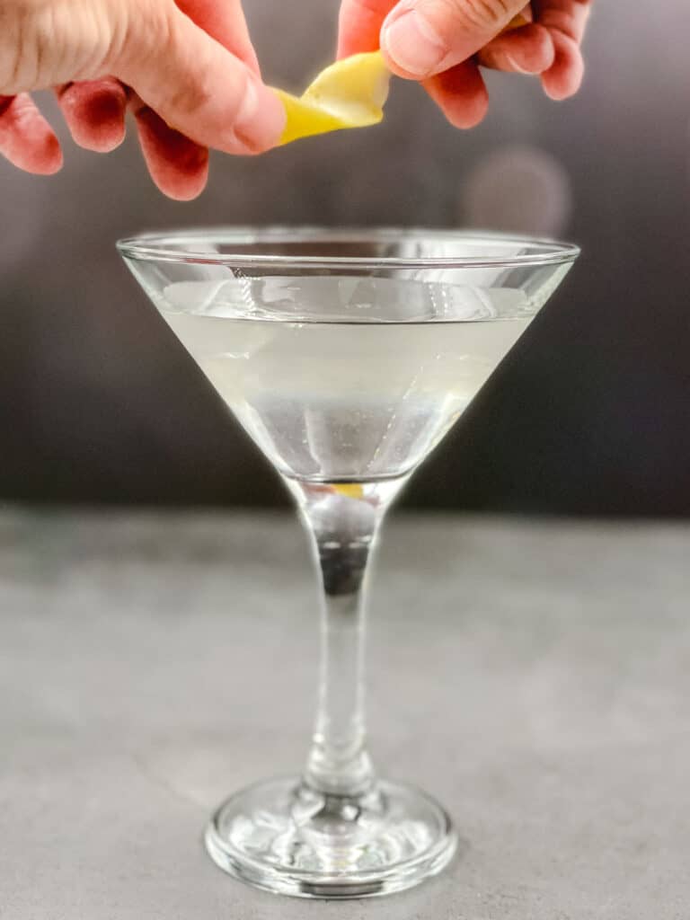 Someone twisting lemon peel above a martini glass.