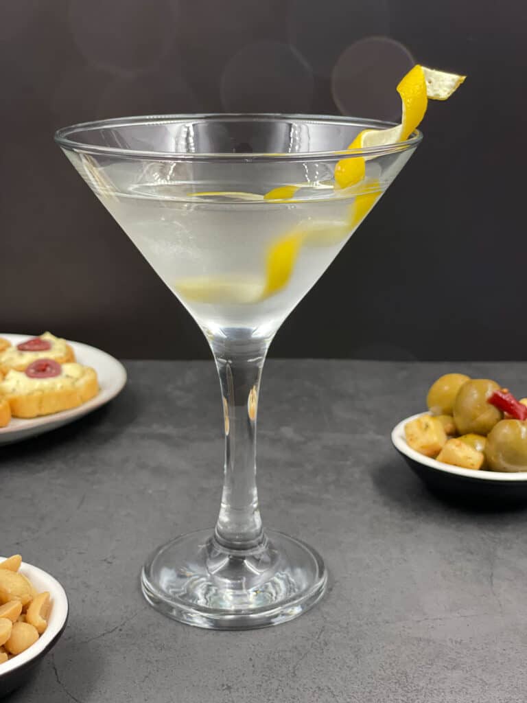 A vodka martini in a cocktail glass with a lemon twist and nuts and olives in bowls.