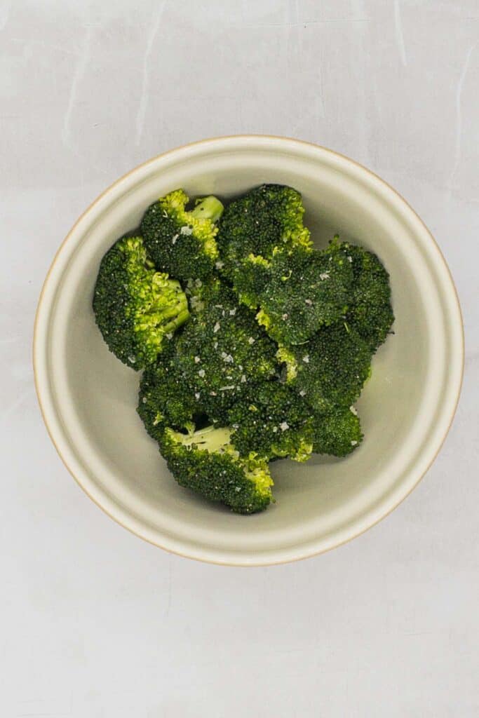 Broccoli in a bowl with Parmesan cheese, oil, salt,, and pepper.