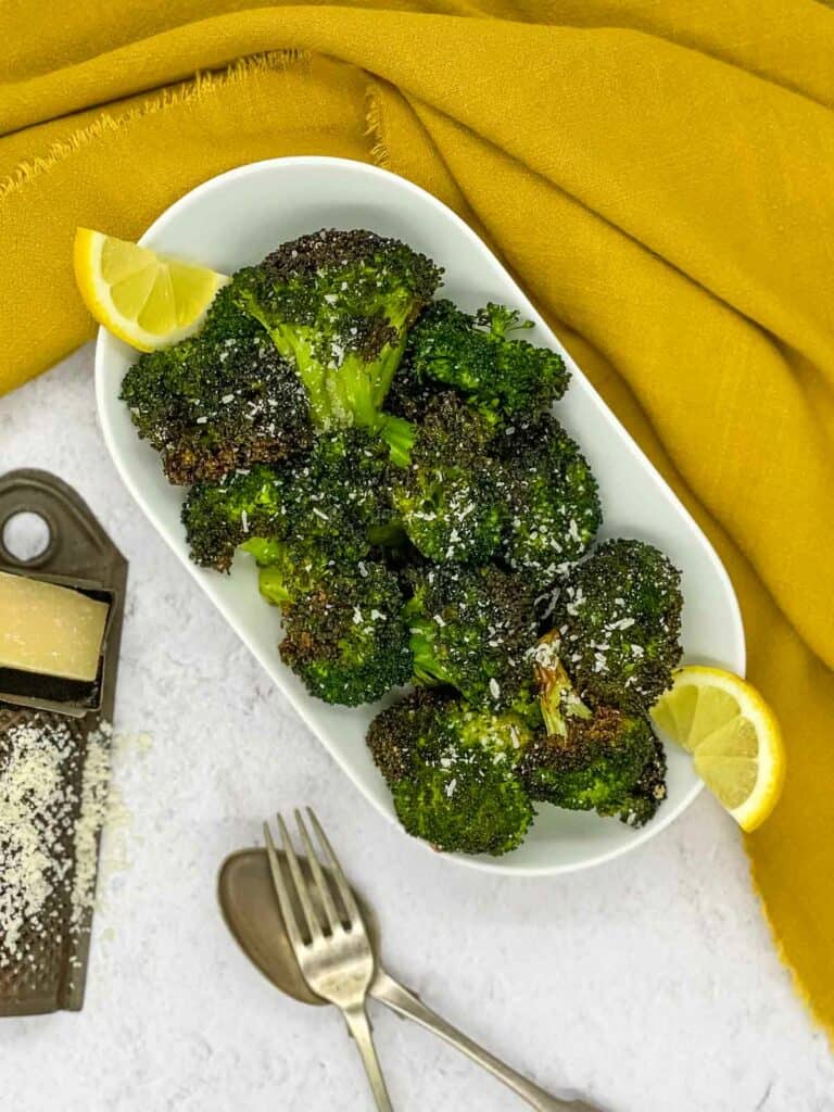 Cooked broccoli on a serving plate, with lemon slices, Parmesan cheese, and a fork, and spoon.