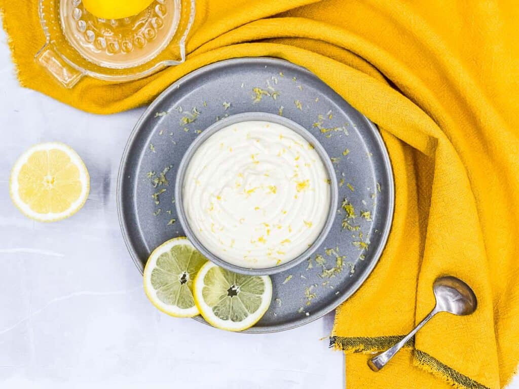 A delicious bowl of lemon aioli on a plate with slices of lemon and a spoon.
