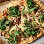 A delightful mushroom pizza adorned with arugula and chicken, sprinkled with herbs, rests on parchment paper next to a bottle of olive oil.