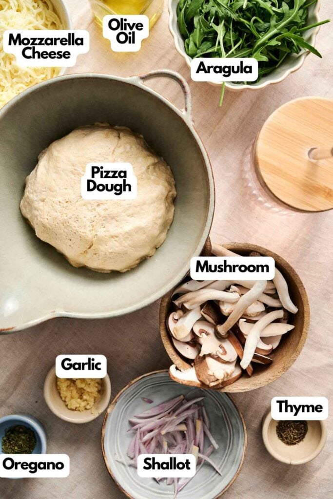 Ingredients for mushroom pizza: a bowl of pizza dough, mozzarella cheese, olive oil, arugula, sliced mushrooms, garlic, shallot, thyme, and oregano on a countertop.