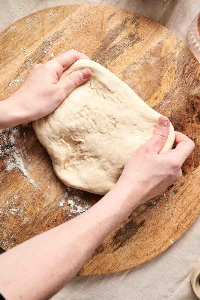 Hands kneading dough on a circular wooden board, with some flour scattered around&mdash;preparing the perfect base for a mouthwatering mushroom pizza.