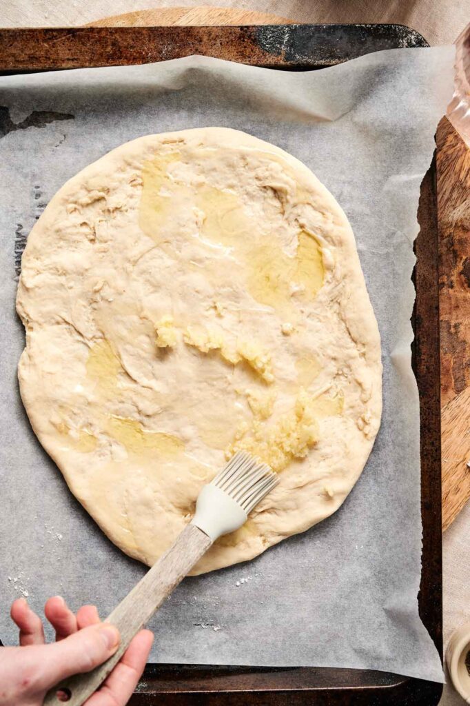 A hand using a brush gently spreads garlic butter over rolled-out pizza dough on a parchment-lined baking tray, preparing the perfect foundation for a delicious mushroom pizza.