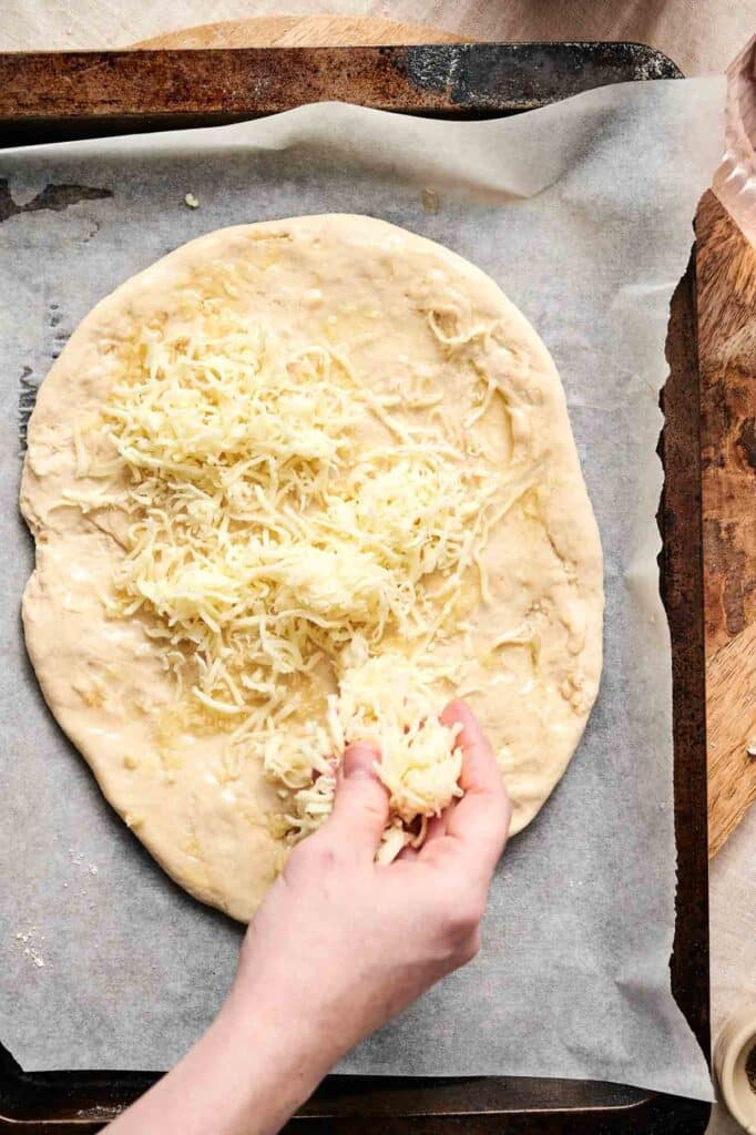 A hand sprinkles shredded cheese onto a round, unbaked mushroom pizza crust resting on parchment paper.