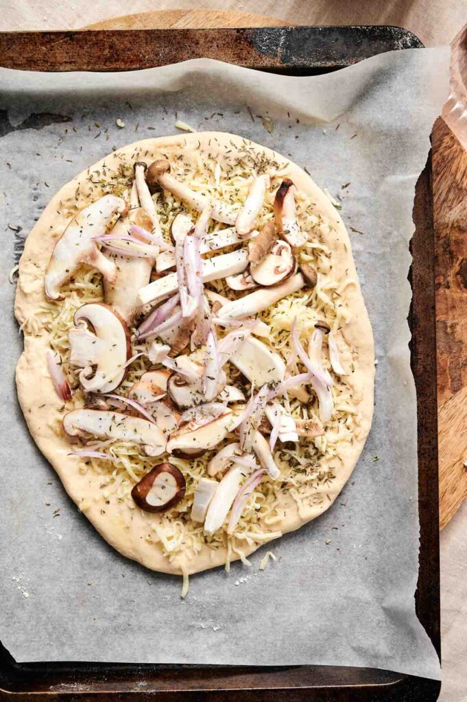 A delicious mushroom pizza awaits, unbaked and ready on a parchment-lined tray. It's topped with fresh onions, melting cheese, and fragrant herbs.