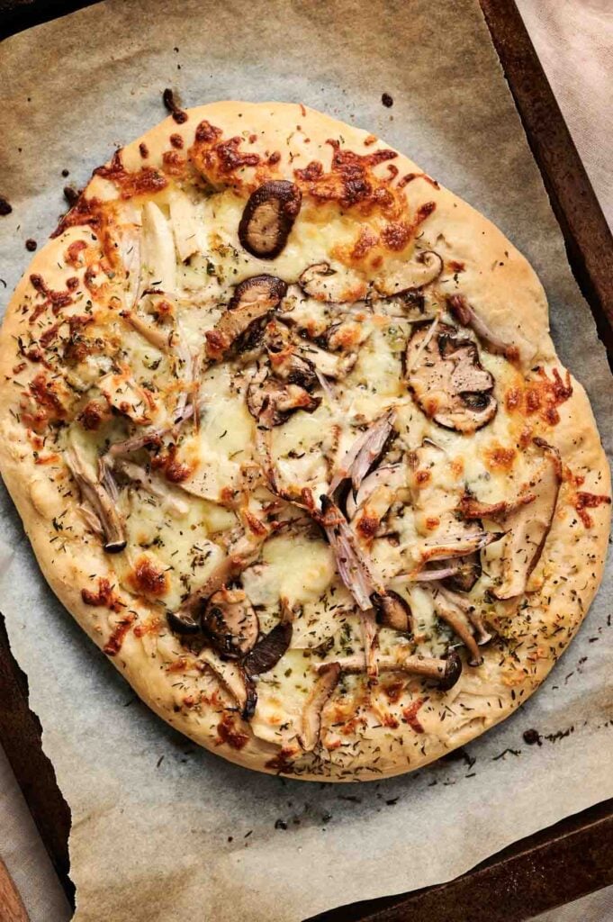Mouthwatering mushroom pizza features an oval shape topped with mushrooms, shredded chicken, melted cheese, and herbs on parchment paper.