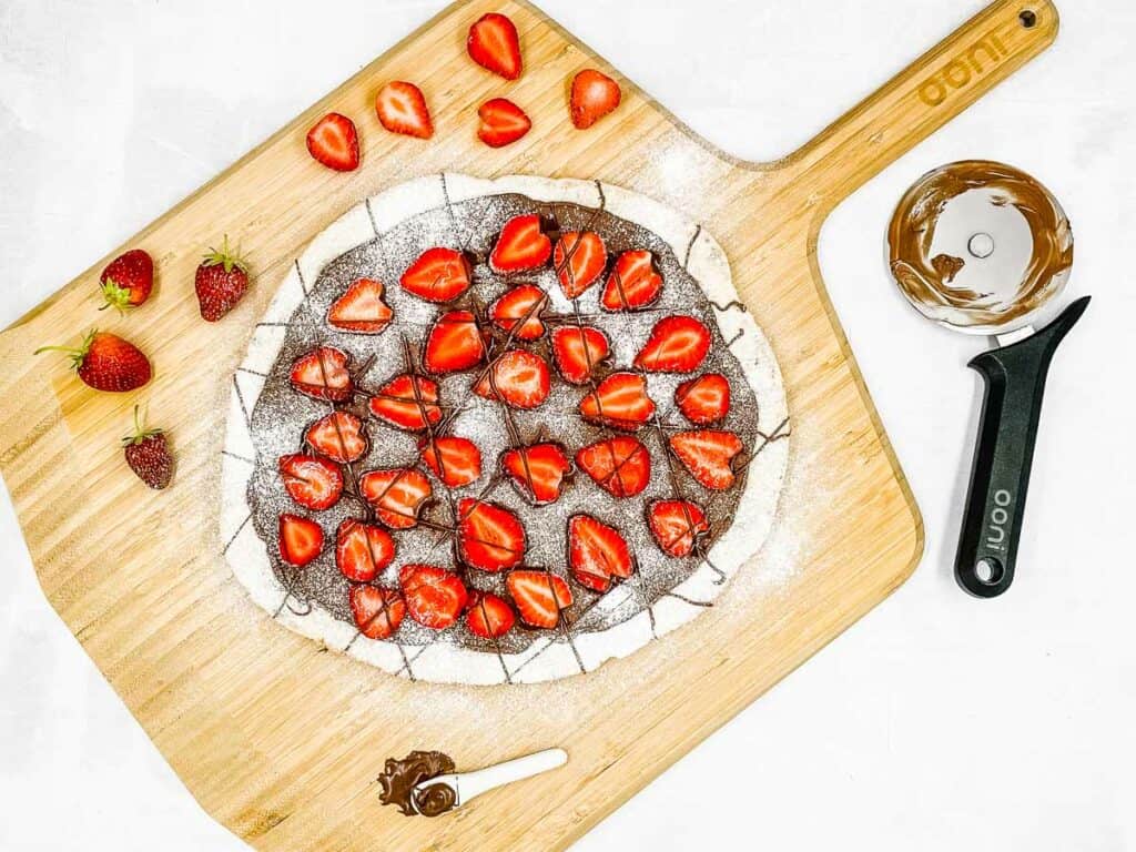 A Nutella dessert pizza on a pizza board with a pizza slicer and strawberries.