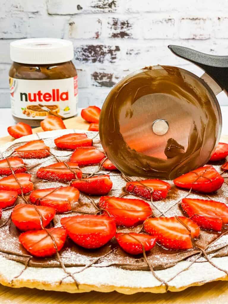 A pizza cutter cutting into a Nutella pizza, with a jar of Nutella in the background.