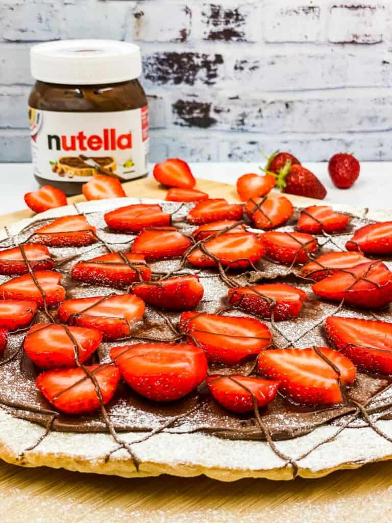 A Nutella pizza with strawberries, with a jar of Nutella chocolate and hazelnut spread in the background.