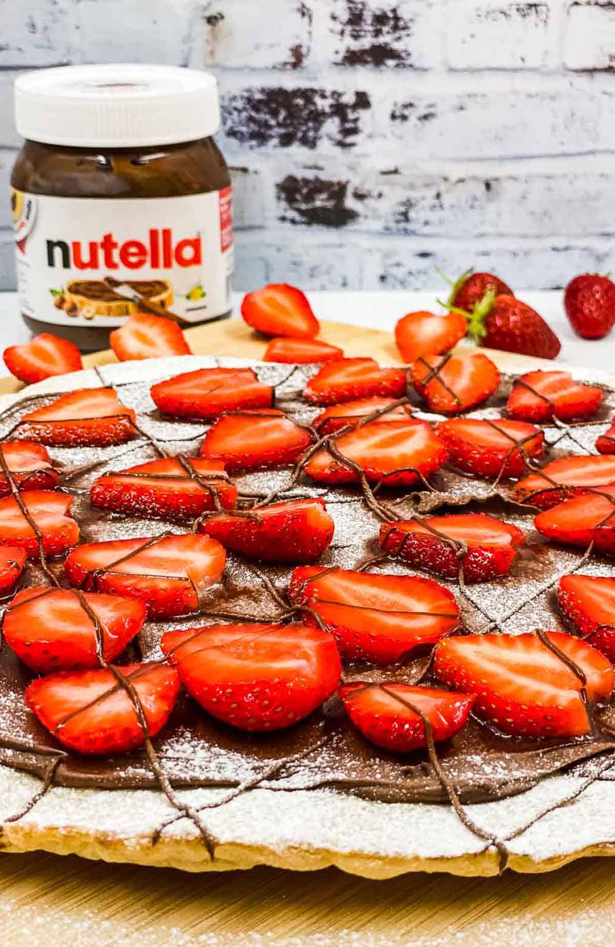 Delicious Nutella pizza with fresh sttrawberries and a jar of Nutella spread in the background.