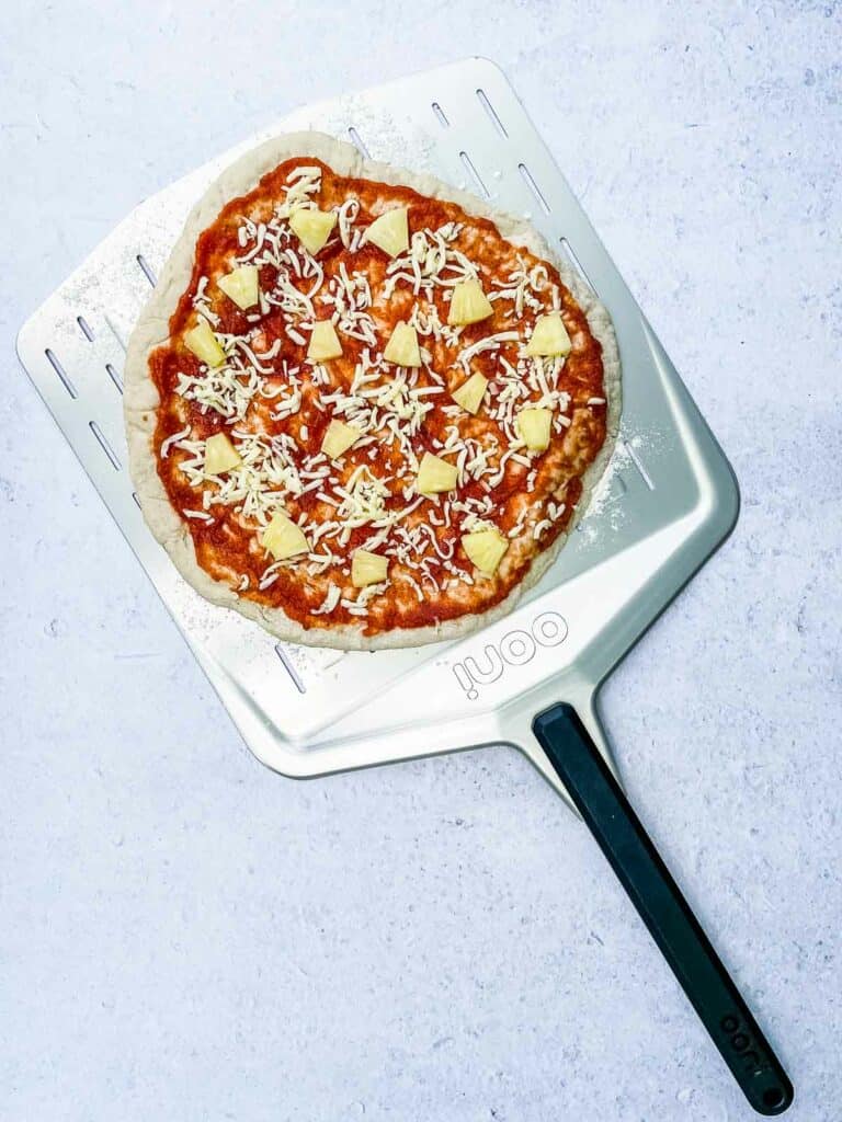 An uncooked pizza crust with pizza sauce, mozzarella cheese, sweet pineapple, peppers, and vegetarian ham.