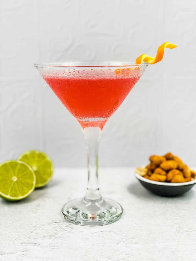 A cosmopolitan cocktail in a martini glass, with limes and nuts around it.