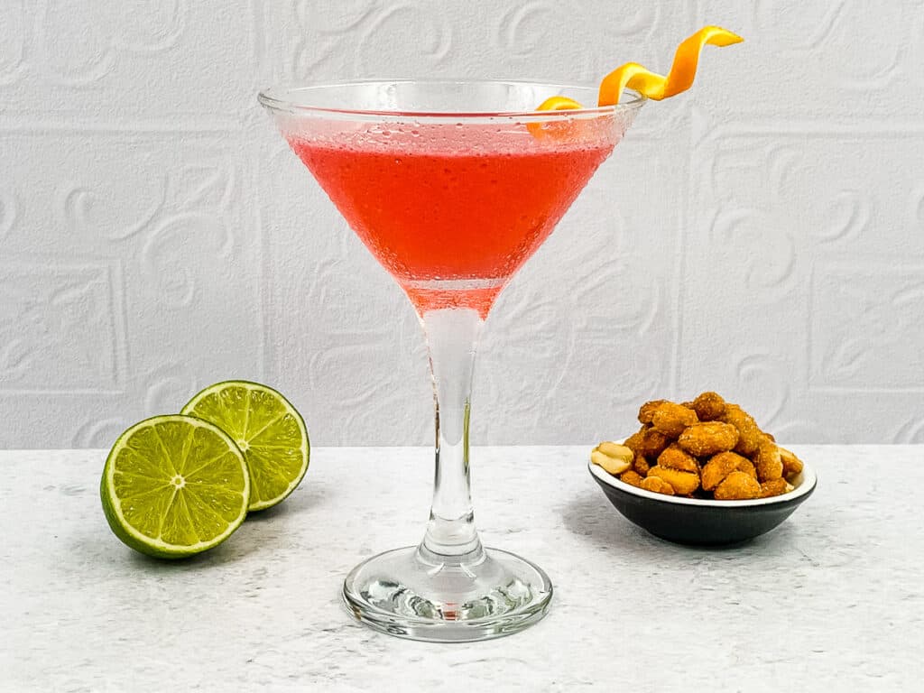 A homemade cosmo drink in a cocktail glass with limes and bar nuts.