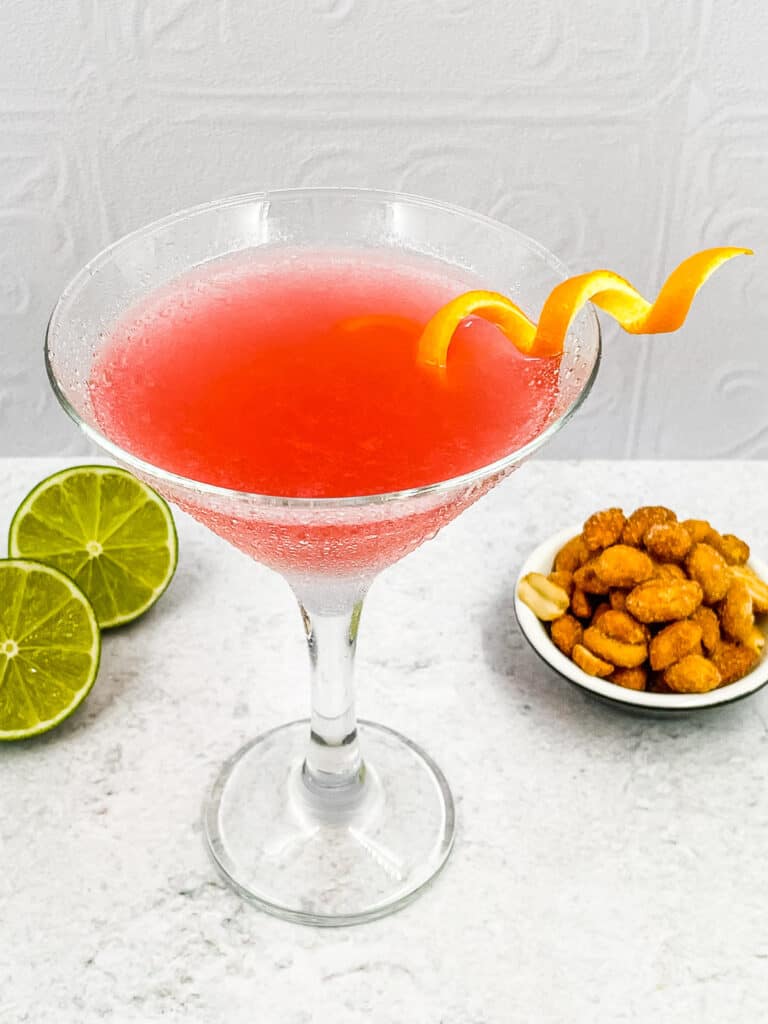 A cosmo cocktail with an orange twist, bar nuts and lime halves.