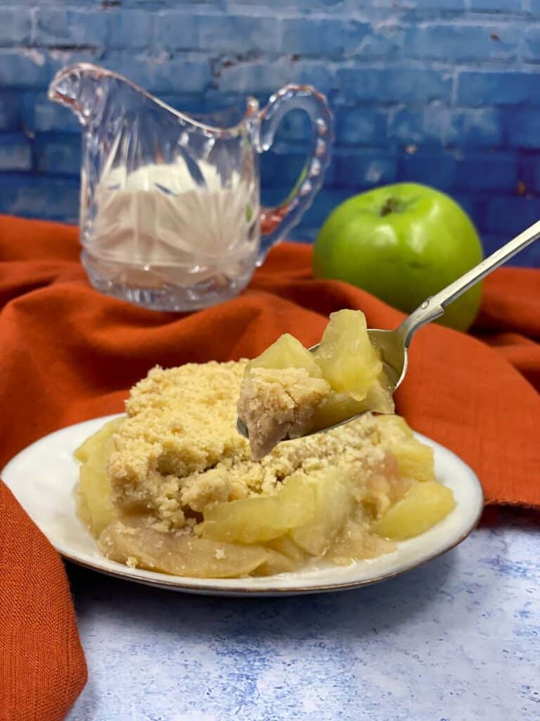 A forkful of gluten free apple crumble and a jug of cream and an apple.