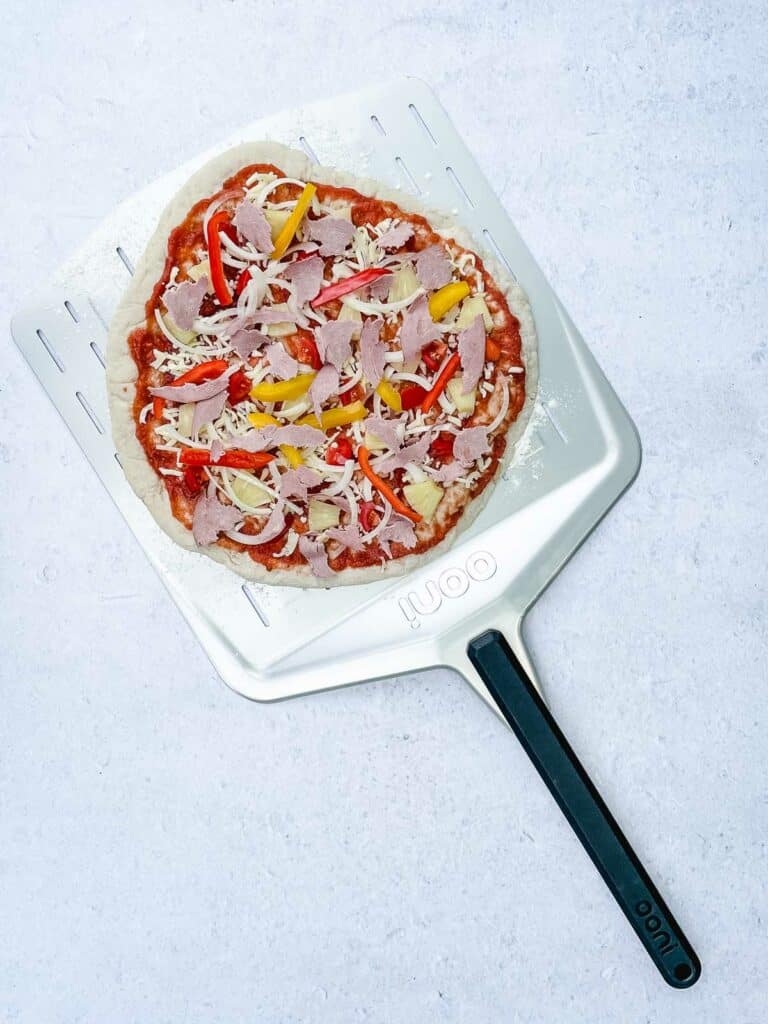 An uncooked pizza crust with pizza sauce, mozzarella cheese, sweet pineapple, peppers, and vegetarian ham.