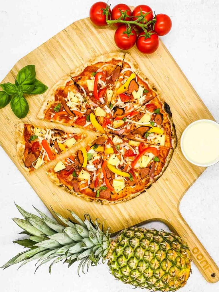 A pineapple pizza on a wooden born with a fresh pineapple, fresh basil leaves, and tomatoes around it.