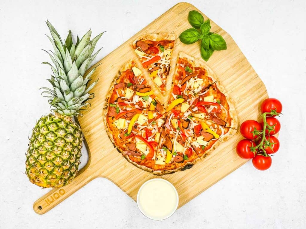 A pineapple pizza, with pineapple, basil, tomatoes, and a pizza dip around it.