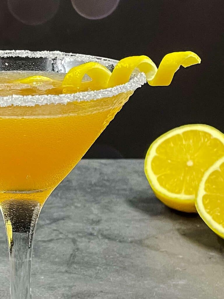 Partial glass of sidecar with a lemon twist and lemons on the surface in the background.