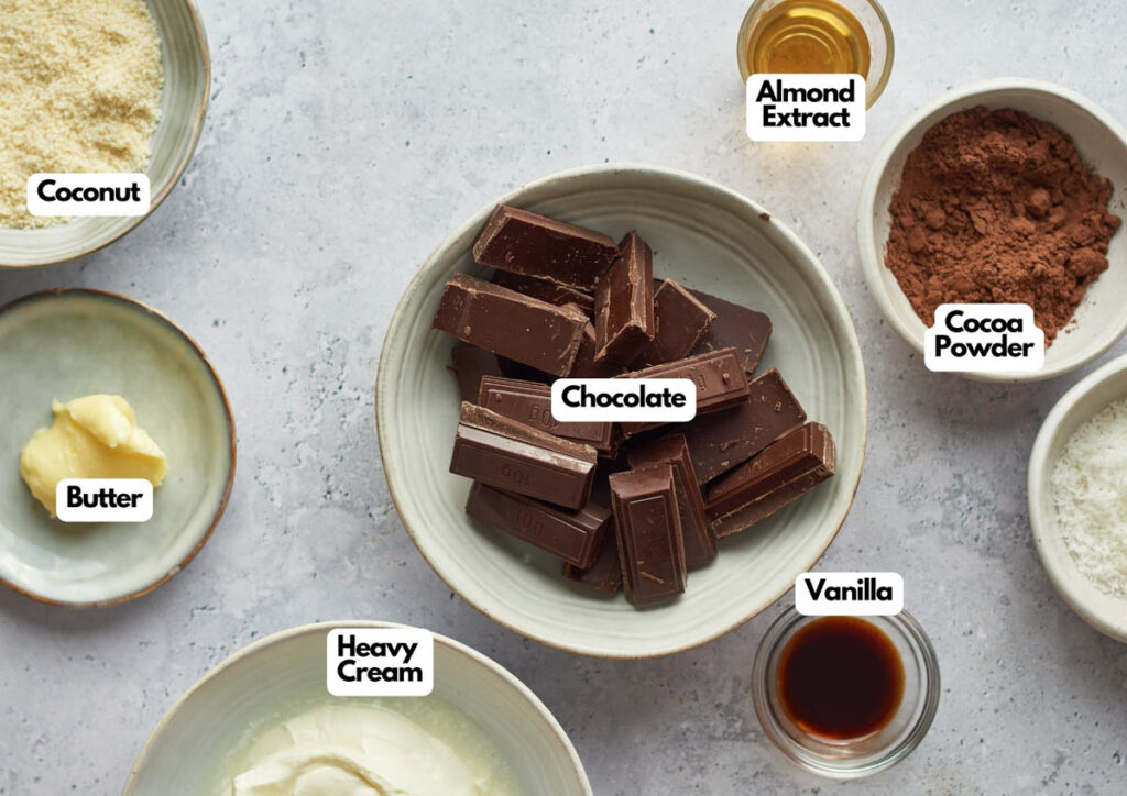 Bowls containing chocolate, almond extract, cocoa powder, vanilla, shredded coconut, heavy cream, and butter&mdash;ingredients perfect for a rich dessert like Baked Alaska&mdash;are arranged on a light surface; each is clearly labeled.